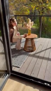 Bej renkli süveter giymiş bir kadının modern terasta kahve içerken, konuşurken ya da video izlerken çekilmiş rahat bir sonbahar yaşam tarzı videosu. Yaşam tarzı vlogları, kahve içeriği, ev dekorasyonu, açık havada yaşam, ya da kişisel hikaye anlatma ve iletişim temaları için mükemmel.