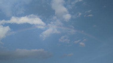 Gökkuşağı soluk mavi bir gökyüzü boyunca uzanır dağınık kabarık bulutlarla huzurlu ve güzel bir doğal sahne yaratır barış ve merak duyguları uyandırır umut ve yeni başlangıçları temsil eder.