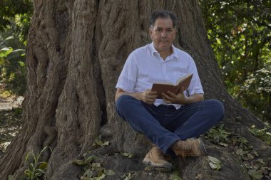 Bir adam yüksek bir ağacın dibinde oturuyor, bir cilt kapağına gömülmüş..