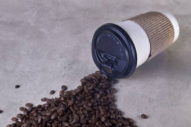 Koyu kapaklı tek kullanımlık bir kahve fincanı açık kumda oturur ve önüne kavrulmuş kahve çekirdeği döker..