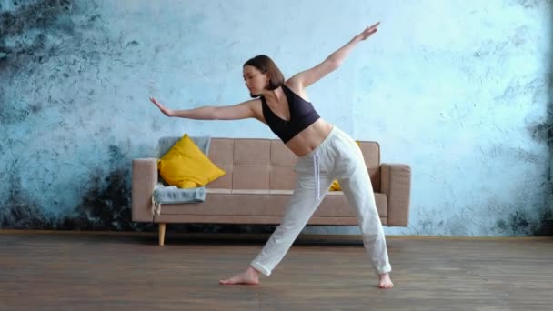 Calma joven morena en blanco hace utthita trikonasana practicando yoga cerca de sofá. — Vídeos de Stock
