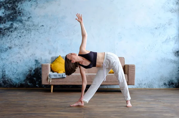 Calma giovane donna bruna in bianco fa utthita trikonasana praticare yoga vicino al divano. — Foto Stock