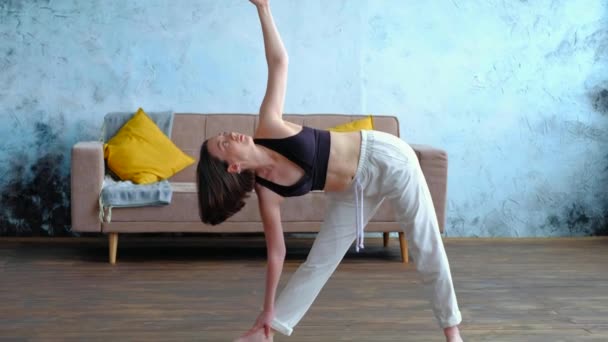 Spokojna młoda kobieta brunetka w bieli robi utthita trikonasana ćwiczy jogę w pobliżu kanapy. — Wideo stockowe