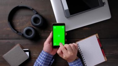 Ahşap masadaki yeşil ekranlı akıllı telefonu kullanan adamın elleri..