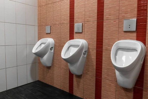 Halka açık bir tuvalette üç beyaz pisuvar fayanslı duvarlar ve karanlık zemin