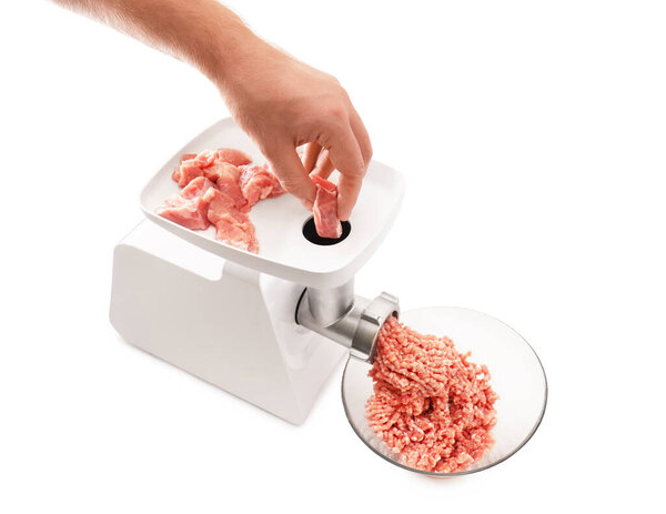 Рука человека толкает сырой кусок свинины в белую электрическую мясорубку, с мясом, собираемым в стеклянную миску. Высокое качество фото
