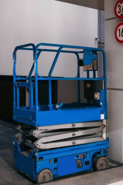 Duvara dayalı mavi bir makas asansörü. Asansör kompakt ve dikey yükselme için tasarlanmış. Hareketlilik için sağlam bir çerçeve ve tekerlek içerir..