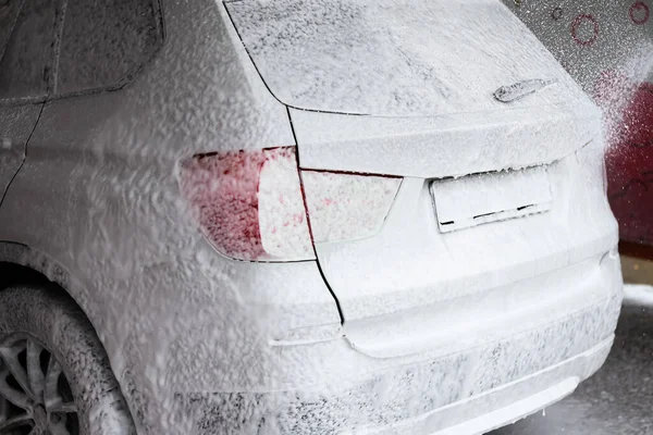 Oto yıkamada köpükle kaplı beyaz bir araba. Köpük, aracın detaylarını gizleyen kalın bir katman oluşturuyor. Ayarlar temiz ve düzenli..