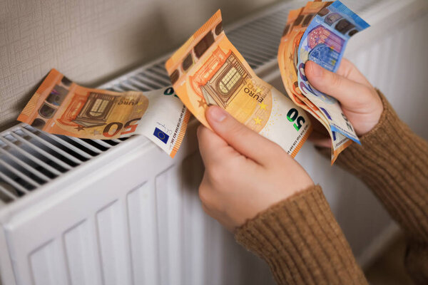 Девушка в свитере считает деньги у радиатора: евро в 50 и 20 номиналов. В комнате холодно, что подчеркивает необходимость обогрева.