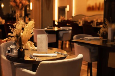 Menü, çatal bıçak takımı ve sıcak ortam ışıkları altında vazoda kurumuş çiçeklerle şık bir restoran masası. Yüksek kalite fotoğraf