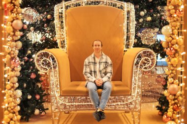 Noel ağaçlarıyla çevrili dev bir tahtta poz veren genç bir adam ve bir alışveriş merkezinde ışıklar. Yüksek kalite fotoğraf