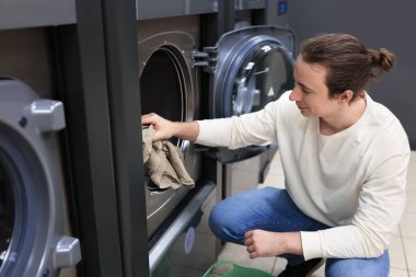 Çamaşırhanedeki çamaşır makinesine çamaşır yüklemek için çömelmiş çömelmiş bir adam. Yüksek kalite fotoğraf