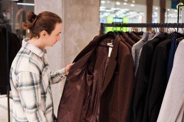 Bun saç stili olan bir adam giyim mağazasında kahverengi palto seçiyor, yeni kıyafet alırken ceket astarını inceliyor. Yüksek kalite fotoğraf