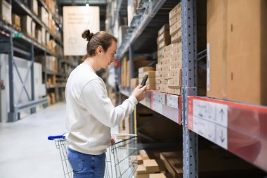 Alış veriş arabasının yanındaki mobilya deposu koridorundaki ürün kodlarını taramak için akıllı telefon kullanan genç adam. Yüksek kalite fotoğraf