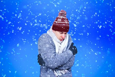 Kışlık şapkalı ve kazaklı dondurucu genç adam mavi arka planda yağan kar altında ısınmak için kendine sarılıyor. Yüksek kalite fotoğraf