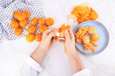 Beyaz mermer bir masada taze mandalinayı soyan erkek ellerinin üst görüntüsü. Tabakta bütün mandalinalar ve soyulmuş bölümler var. Yüksek kalite fotoğraf