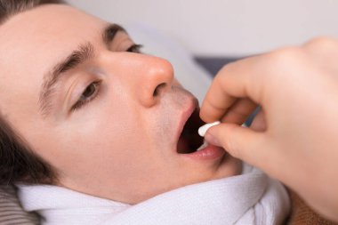 Close up of sick young man with scarf opening mouth to take a white pill from another person's hand. High quality photo
