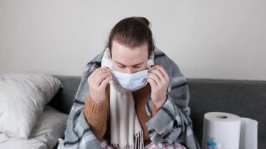 Kanepede otururken koruyucu maske takan atkı ve battaniyeli hasta bir adam. Grip, virüs, karantina ve enfeksiyonun evde yayılmasını önlemek. Yüksek kalite 4k görüntü