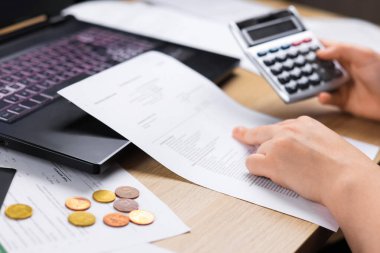 Hesap makinesi kullanan bir kadının ellerinin yakın çekimi ve bir dizüstü bilgisayar ve bozuk paraların yanındaki belgeleri incelemesi. Konsept olarak, ev bütçesi, mali stres, kabarık faturalar veya muhasebe anlamına gelir. Yüksek kalite fotoğraf