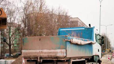 Şehir caddesinde bir kazıcıyla kum taşıyan bir çöp kamyonunun 4K video görüntüleri, bir inşaat alanı, bulutlu bir havada toprak ve malzeme taşıma.