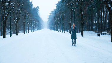 Gri ceketli bir adam, karlı bir orman yolunda yürüyor, soğukta ellerini ısıtıyor. Atmosferik kış parkı sahnesi ağaçlarla kaplı, sisli bir patika. Yüksek kalite fotoğraf