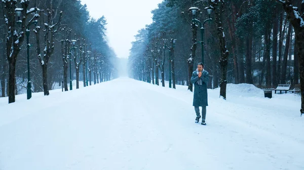 Gri ceketli bir adam, karlı bir orman yolunda yürüyor, soğukta ellerini ısıtıyor. Atmosferik kış parkı sahnesi ağaçlarla kaplı, sisli bir patika. Yüksek kalite fotoğraf