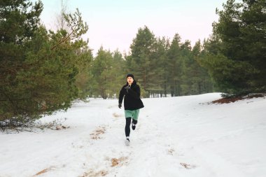 Genç adam karlı bir orman yolunda koşuyor. Sıcak spor giyimli erkek sporcu kış antrenmanı sırasında kameraya doğru koşuyor. Yüksek kalite fotoğraf