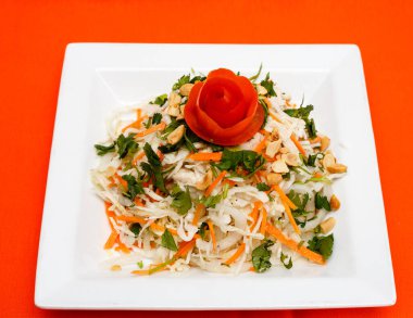 Fıstık ezmesi, kişniş ve domates gülü garnitürle kaplı taze bir Asya lahana salatası, canlı turuncu arka planda beyaz bir tabakta servis edilir. Temiz yemek ve mutfak temaları için ideal.