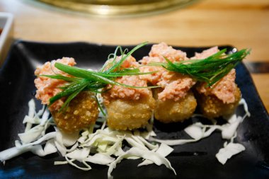 Üstü taze, baharatlı somon tartarıyla kaplanmış, koyu bir tabakta ince dilimlenmiş soğanla süslenmiş, altın kızartılmış suşi rulolarından oluşan gurme aperatifi..