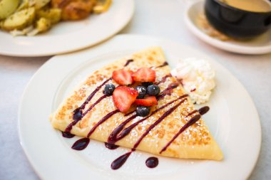 Çikolata şurubuyla serpilmiş tatlı krepi ve üstüne taze çilek, yaban mersini ve bir kafe ortamında dikey olarak çekilmiş bir parça krema..