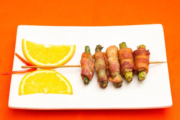 Bir şişte beş pastırmaya sarılmış kuşkonmaz mızrağının beyaz bir tabakta parlak portakal dilimleriyle servis edildiği makro bir stüdyo çekimi. Aperatifler, gurme atıştırmalıklar ve makro gıda ayrıntıları için idealdir..