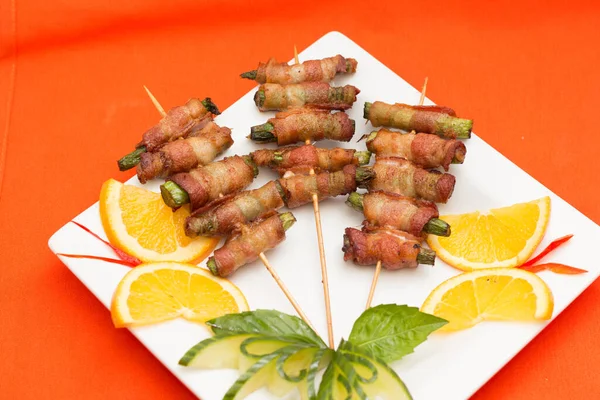 Portakal dilimleri, salatalık ve fesleğen garnitürlü beyaz bir tabağa yerleştirilmiş çoklu pastırmaya sarılmış kuşkonmaz şişlerinden oluşan geniş bir stüdyo çekimi. Yemek servisi, aperatifler ve parti yemekleri için uygun..