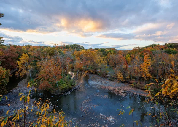 Sonbahar renklerinde, turuncu ve sarı ağaçlarla dolu bir nehir vadisinin panoramik manzarası, dramatik bir gökyüzü altında, yumuşak günbatımı bulutları. Önplan yapraklarıyla çerçevelenmiş.