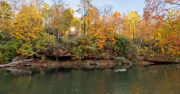 Sakin bir nehrin geniş panoramik HDR manzarası kıyılarındaki canlı sarı ve turuncu sonbahar ağaçlarını yansıtır. Bir güneş yıldızı kayalıklardaki küçük tahta gözetleme kulesini delip geçer..