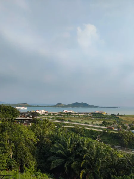 Yeşil tepeler, tropik bitki örtüsü ve bulutlu bir gökyüzünün altında sakin bir deniz manzarası.
