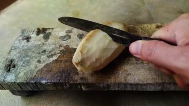 Yakından çekilmiş bir fotoğraf, beton bir yüzey üzerinde bıçakla cassava (yucca kökü) soyan birini gösteriyor..