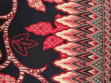 Geleneksel bir batik kumaş tasarımı. Kırmızı ve krem yaprak desenleri var. Siyah arka planda karmaşık detaylar var..
