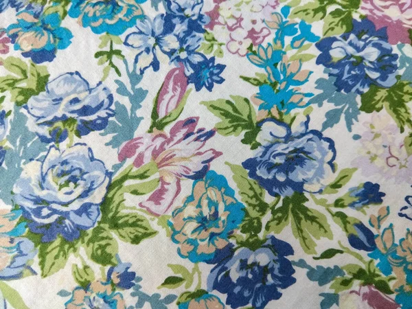 Beyaz arka planda yeşil yapraklı mavi, mor ve pembe çiçeklerden oluşan renkli bir kumaş deseni.