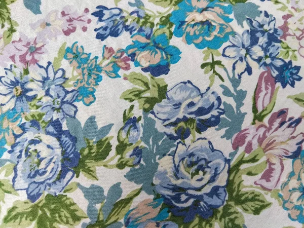 Beyaz arka planda yeşil yapraklı mavi, mor ve pembe çiçeklerden oluşan renkli bir kumaş deseni.