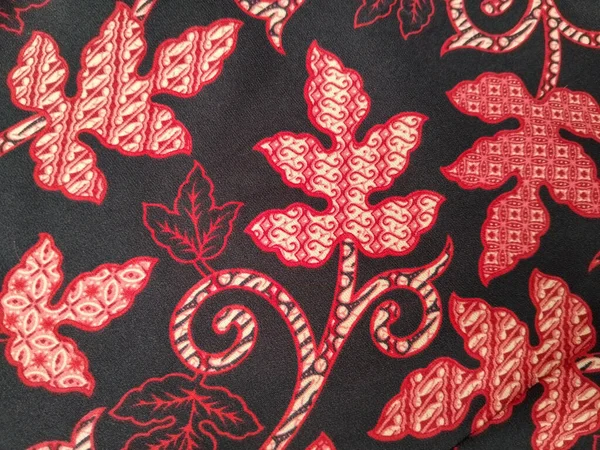 Geleneksel bir batik kumaş tasarımı. Kırmızı ve krem yaprak desenleri var. Siyah arka planda karmaşık detaylar var..