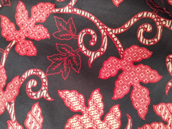 Geleneksel bir batik kumaş tasarımı. Kırmızı ve krem yaprak desenleri var. Siyah arka planda karmaşık detaylar var..
