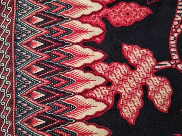 Geleneksel bir batik kumaş tasarımı. Kırmızı ve krem yaprak desenleri var. Siyah arka planda karmaşık detaylar var..