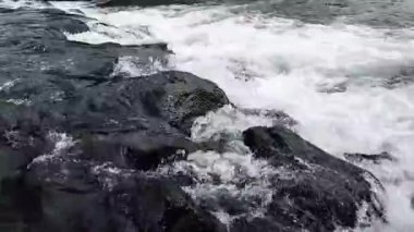 Nehir suyunun akışını yakından görmek dinamik dalgalanmalar yaratır ve karanlık kayaların üzerinde hareket ederken su sıçratır.