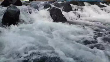 Nehir suyunun akışını yakından görmek dinamik dalgalanmalar yaratır ve karanlık kayaların üzerinde hareket ederken su sıçratır.