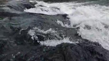 Nehir suyunun akışını yakından görmek dinamik dalgalanmalar yaratır ve karanlık kayaların üzerinde hareket ederken su sıçratır.