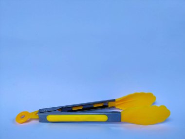 Açık mavi arka plana yerleştirilmiş paslanmaz çelik ve silikondan yapılmış bir çift sarı mutfak maşası.