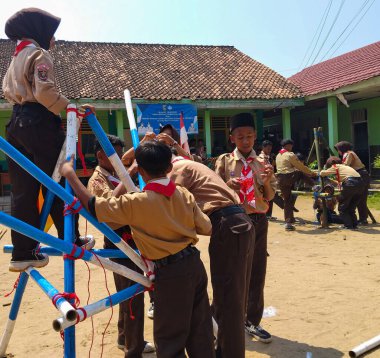 Bir grup Endonezyalı izci, kırmızı iplerle bağlanmış mavi ve beyaz direklerle bir yapı inşa etmek için açık havada birlikte çalışıyorlar.