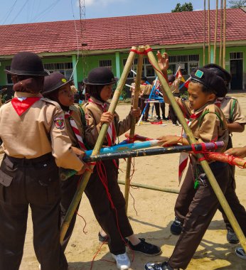 Bir grup Endonezyalı izci, kırmızı iplerle bağlanmış mavi ve beyaz direklerle bir yapı inşa etmek için açık havada birlikte çalışıyorlar.