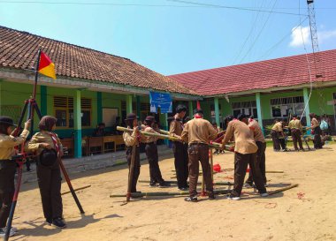 Bir grup Endonezyalı izci, kırmızı iplerle bağlanmış mavi ve beyaz direklerle bir yapı inşa etmek için açık havada birlikte çalışıyorlar.