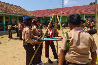 Bir grup Endonezyalı izci, kırmızı iplerle bağlanmış mavi ve beyaz direklerle bir yapı inşa etmek için açık havada birlikte çalışıyorlar.